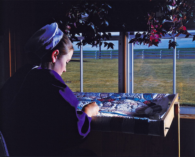 Shows an Amish girl hand quilting. Lancaster County Amish girl hand quilting at a window.